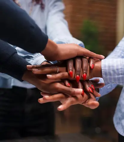 A close-up image of four hands stacked on top of each other. The hands are all dark-skinned and the person on top has red nail polish. The image conveys a sense of unity and teamwork.